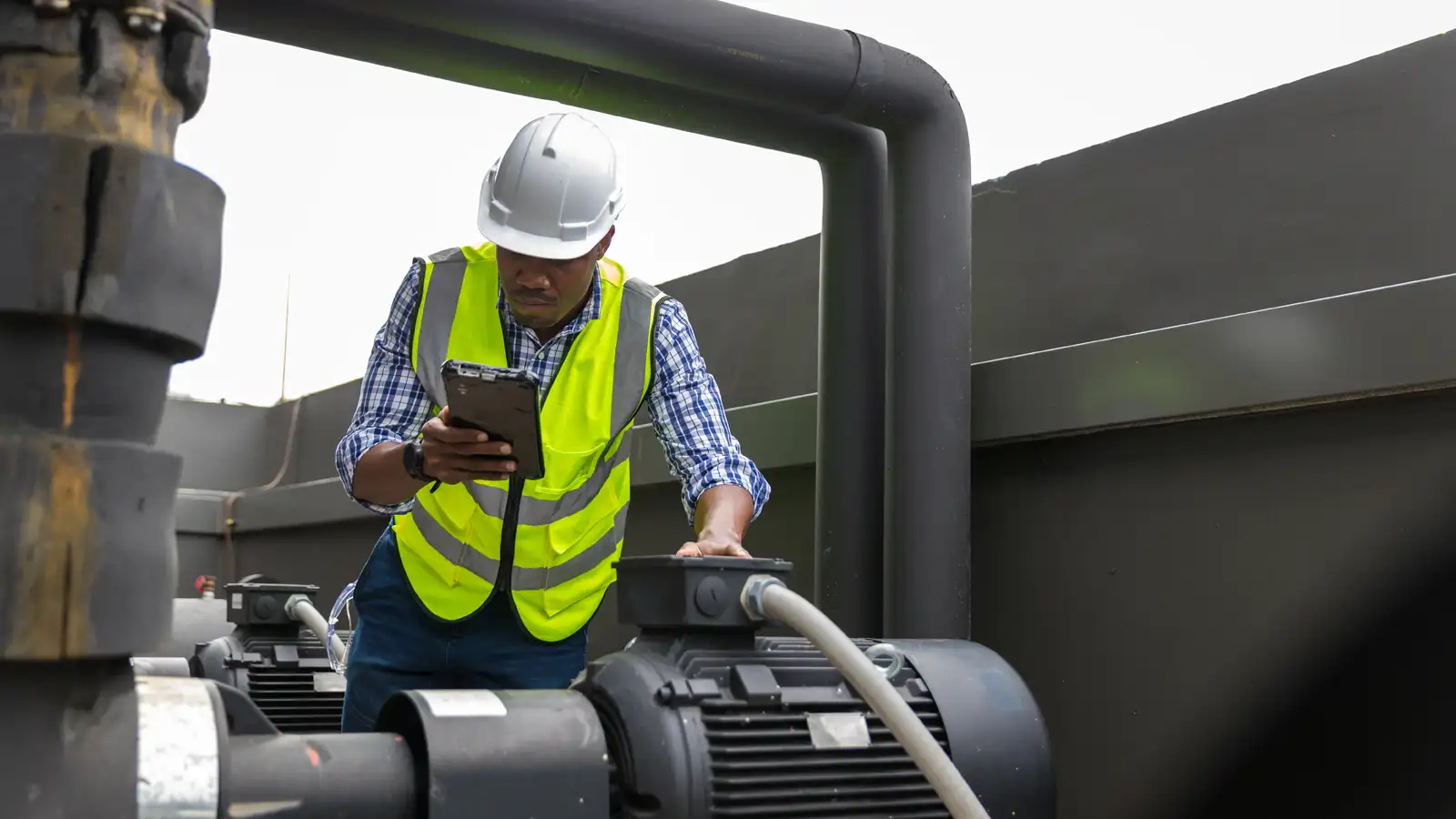 Technician in a hard hat and hi-vis vest inspecting industrial pumps on a rugged tablet during a smart monitoring check.